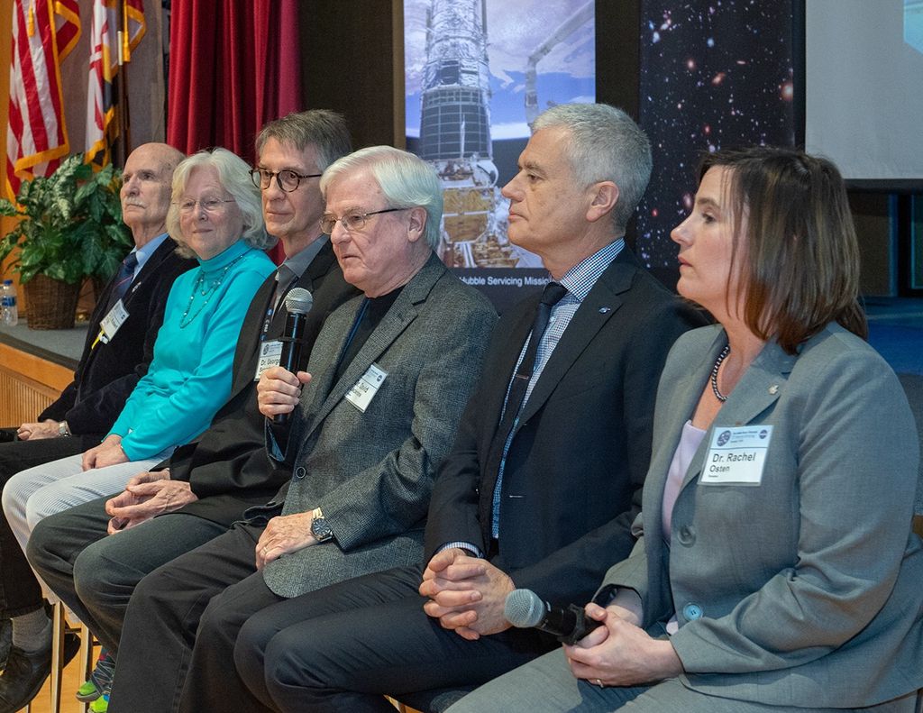 Four men and two women sit in a row in chairs in front of the stage, with Osten (far-right) closest to the camera and O'Dell (far left) farthest from it.