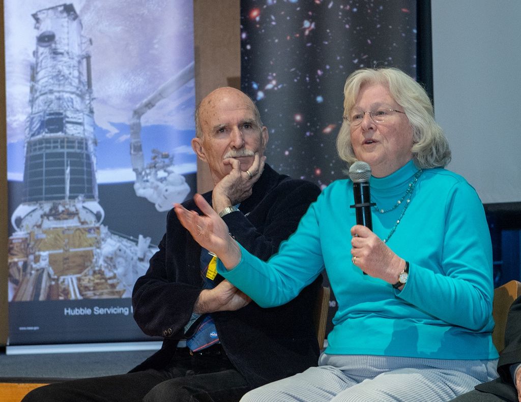 Sara Heap (right) speaks to the audience through the mic in her left hand and motions with her right hand, while C. Robert O'Dell (left) listens.