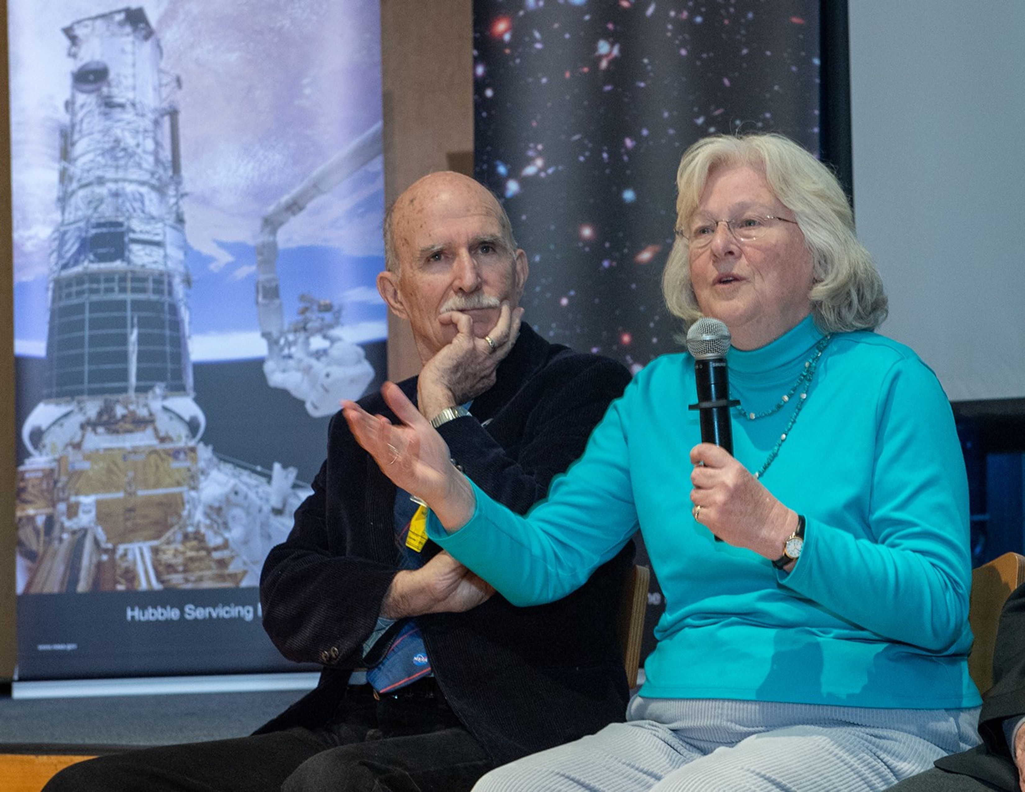 Sara Heap (right) speaks to the audience through the mic in her left hand and motions with her right hand, while C. Robert O'Dell (left) listens.