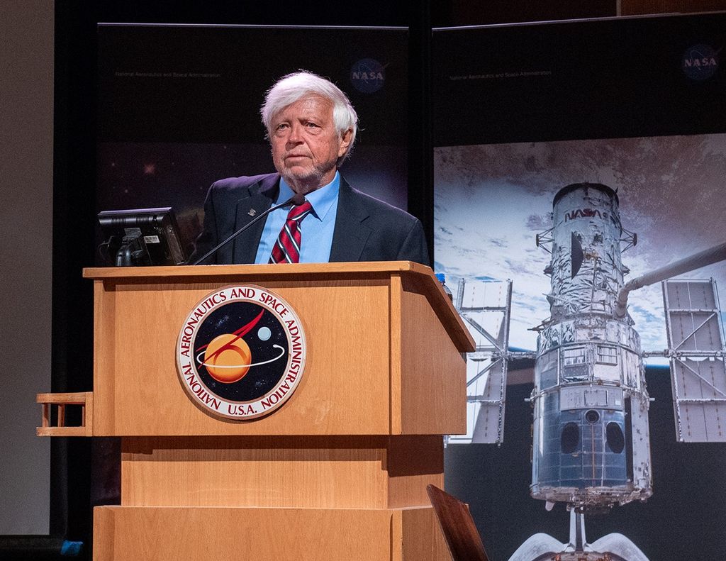 Edward Weiler stands at the podium addressing the audience, with a banner of the Hubble Space Telescope over the Earth behind him.