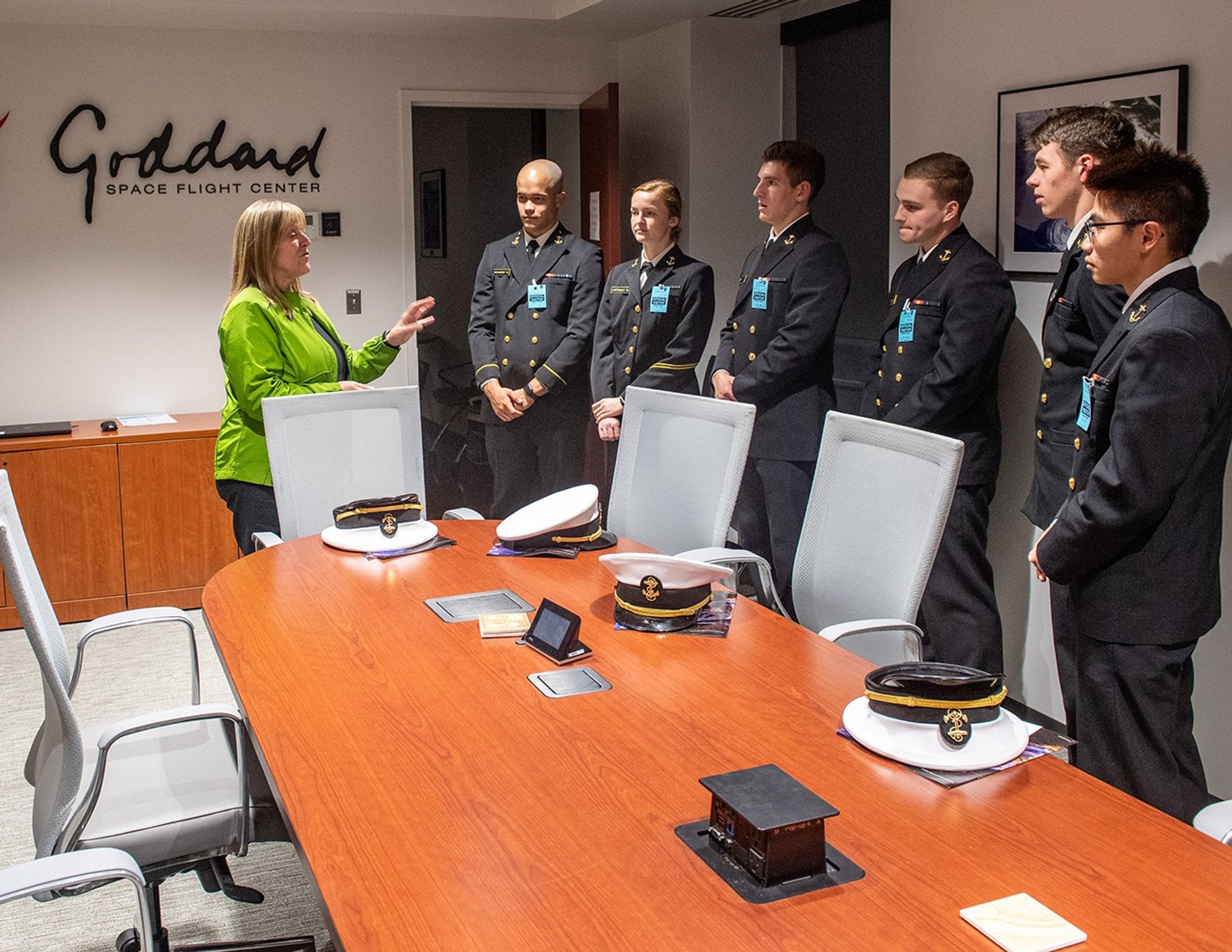 Kay Hire, left, speaks to a group of Midshipmen (five men and one woman are visible in this image), right, around a conference room table.