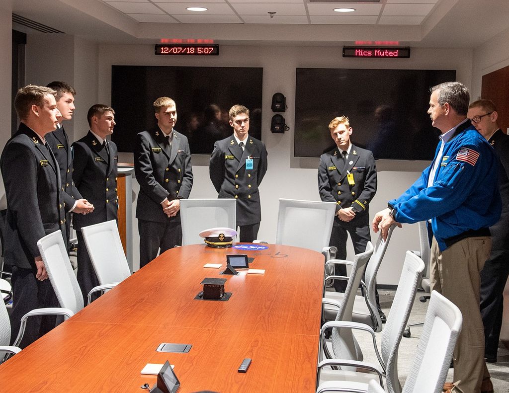 Scott Altman, right, speaks to a group of Navy midshipmen, left (seven men are visible in the image), around a conference room table.