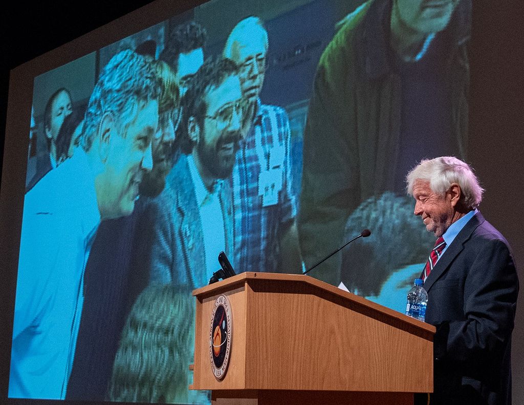 Ed Weiler addresses the crowd while, in the background, a slide shows a picture of several people smiling as they gather around a computer.