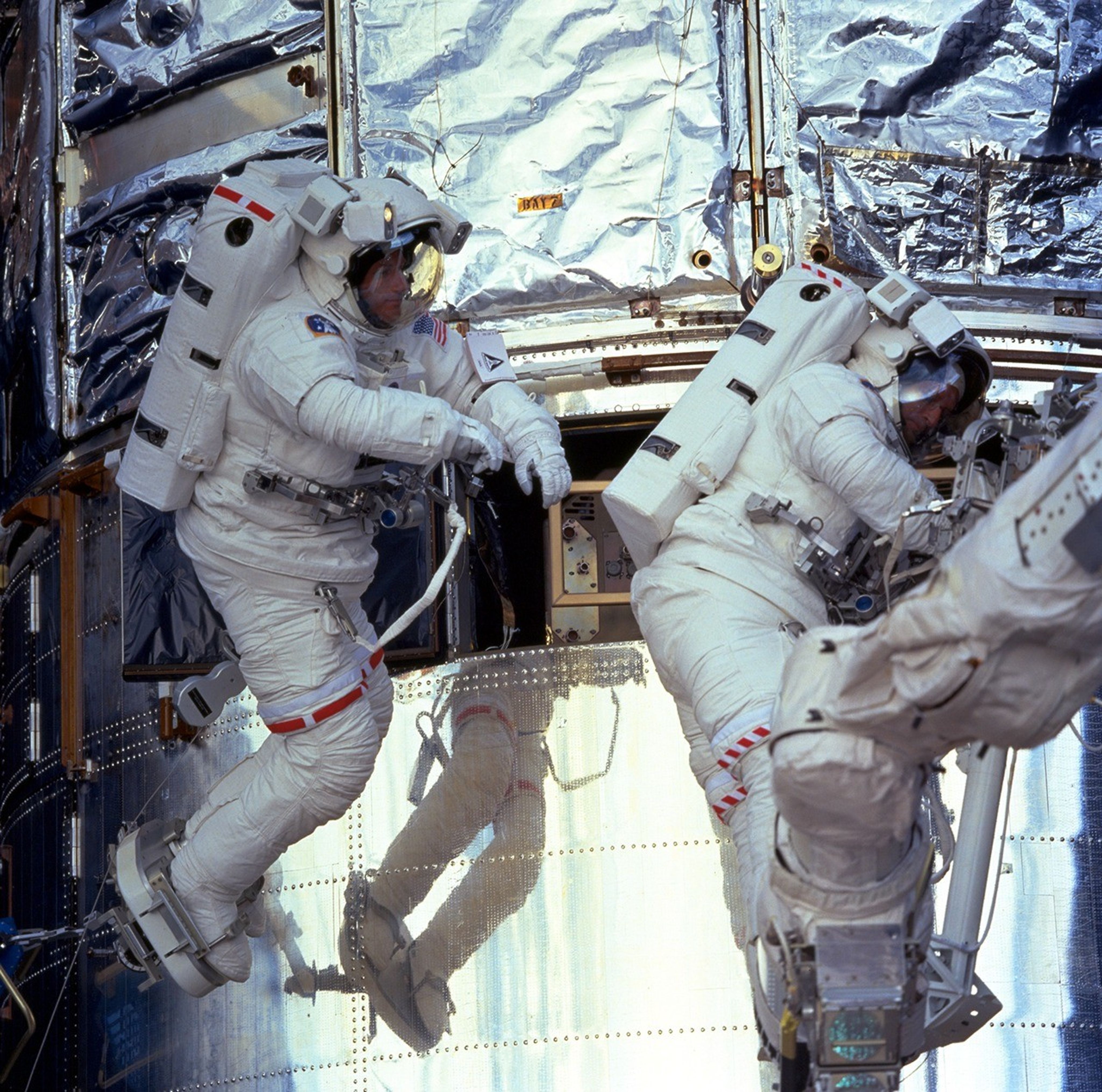 Two astronauts on a space walk in front of the Hubble Space Telescope. One of Hubble's instrument bays is open.