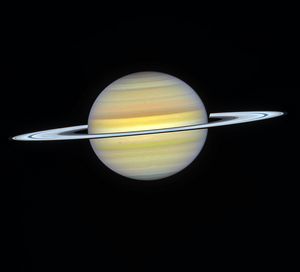 Colorful stripes in yellow, white, and reddish-orange cover the planet. Saturn is tilted slightly toward us, allowing the Sun to illuminate the top of its rings. Three of Saturn's moons are visible as faint white dots: Janus, just below its rings on the left; Epimetheus, to the right of its rings on the right side; and Mimas, which is directly in front of Saturn, slightly below its rings, on the left side of the planet, casting a shadow on its clouds.