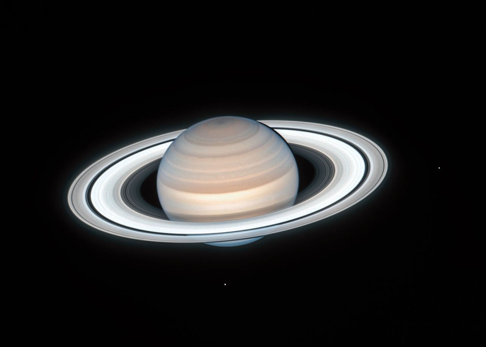 Image of the ringed planet Saturn, with two of its moons visible as tiny dots, one to the right of the planet and one below it.