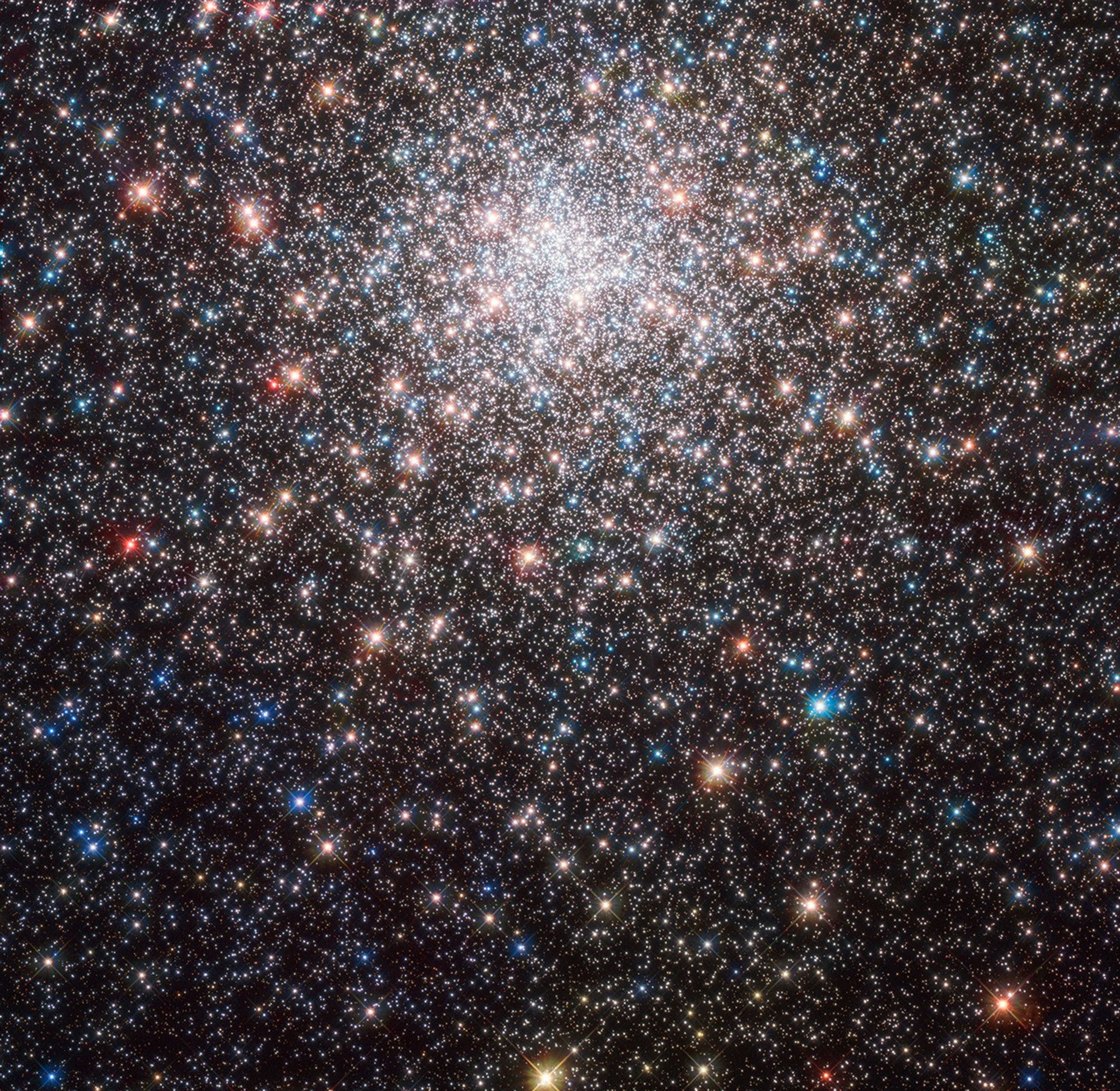 A sphere of stars fills the upper half of the image. The sphere is cut off at the top but extends through out the rest of the image. Stars in bright-white, blue, orange, red, and yellow highlight this colorful globular cluster.