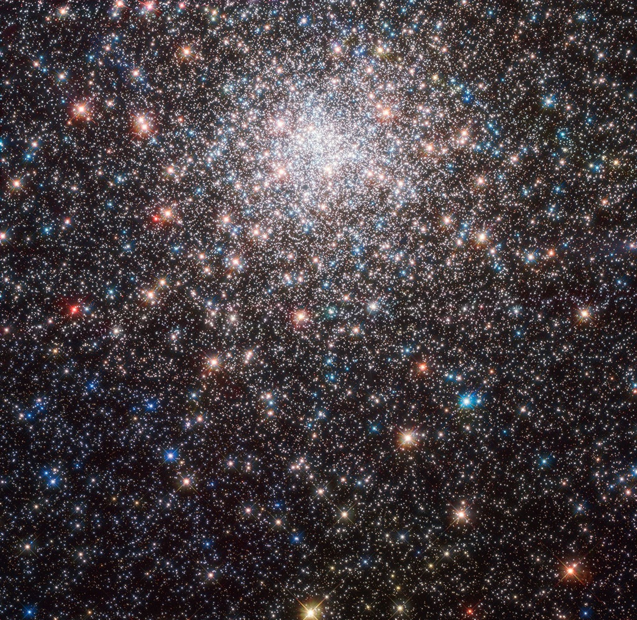 A sphere of stars fills the upper half of the image. The sphere is cut off at the top but extends through out the rest of the image. Stars in bright-white, blue, orange, red, and yellow highlight this colorful globular cluster.