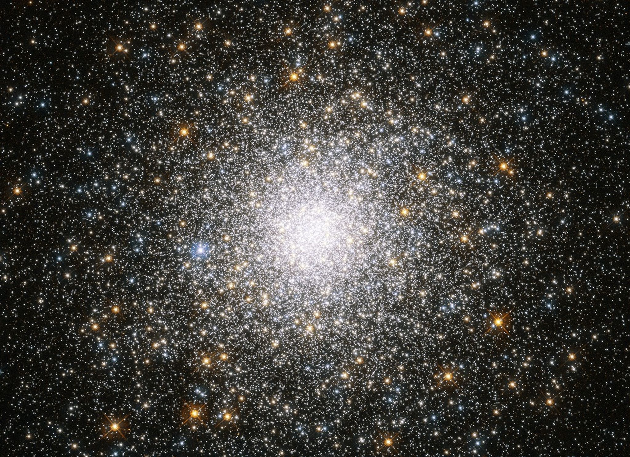 A splotch of stars sits at the center of the image. Its bright-white core is filled with stars. White, yellow-orange, and blue stars dot the view beyond the bright core.