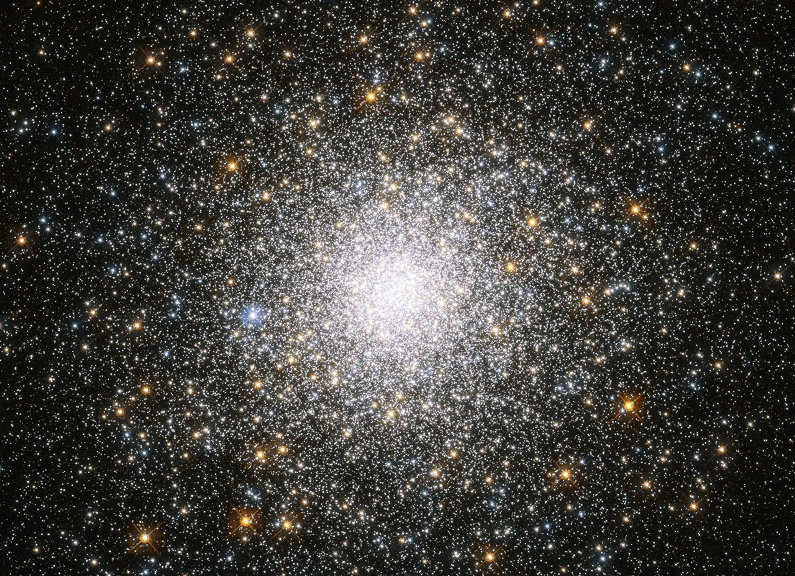 A splotch of stars sits at the center of the image. Its bright-white core is filled with stars. White, yellow-orange, and blue stars dot the view beyond the bright core.