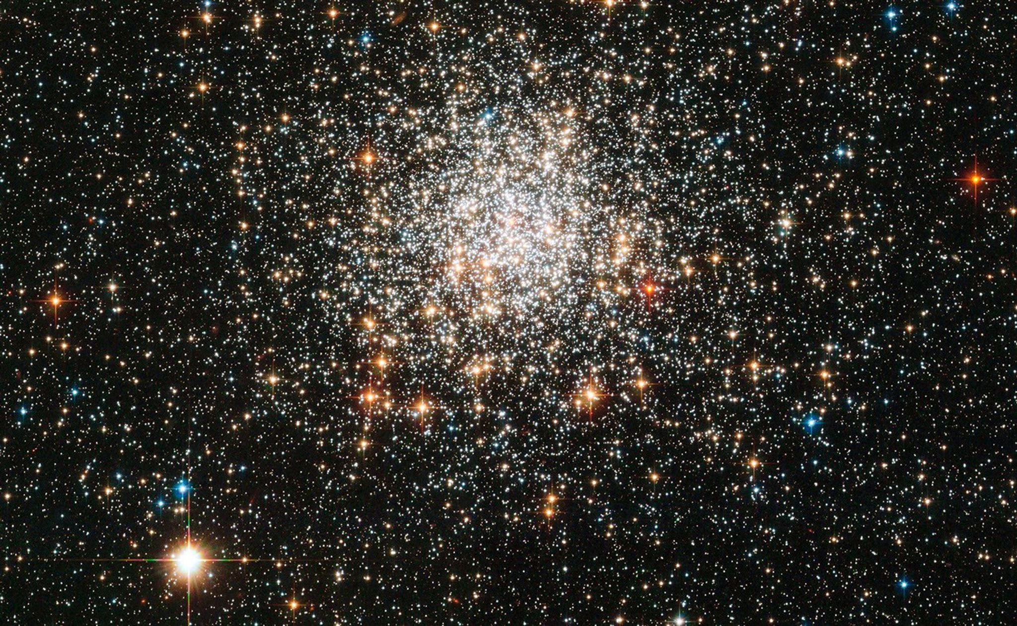 White, red-orange, and a few blue stars packed tightly near the center in a sphere-shape, and thinning out farther from the center.