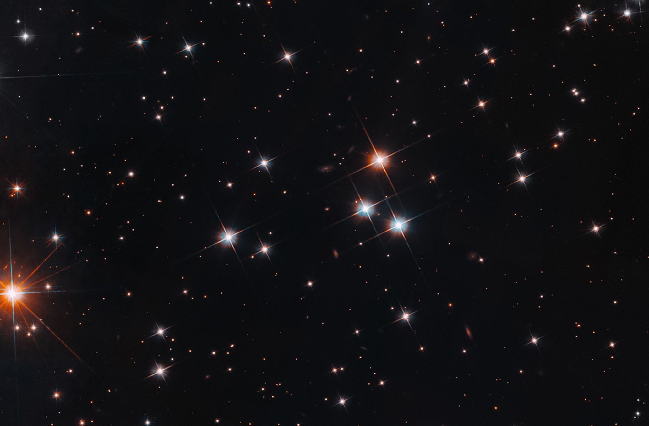 A scattering of stars shine against the black background of space. Most of the stars are white, but some are red. Some small, dim background galaxies are also visible.