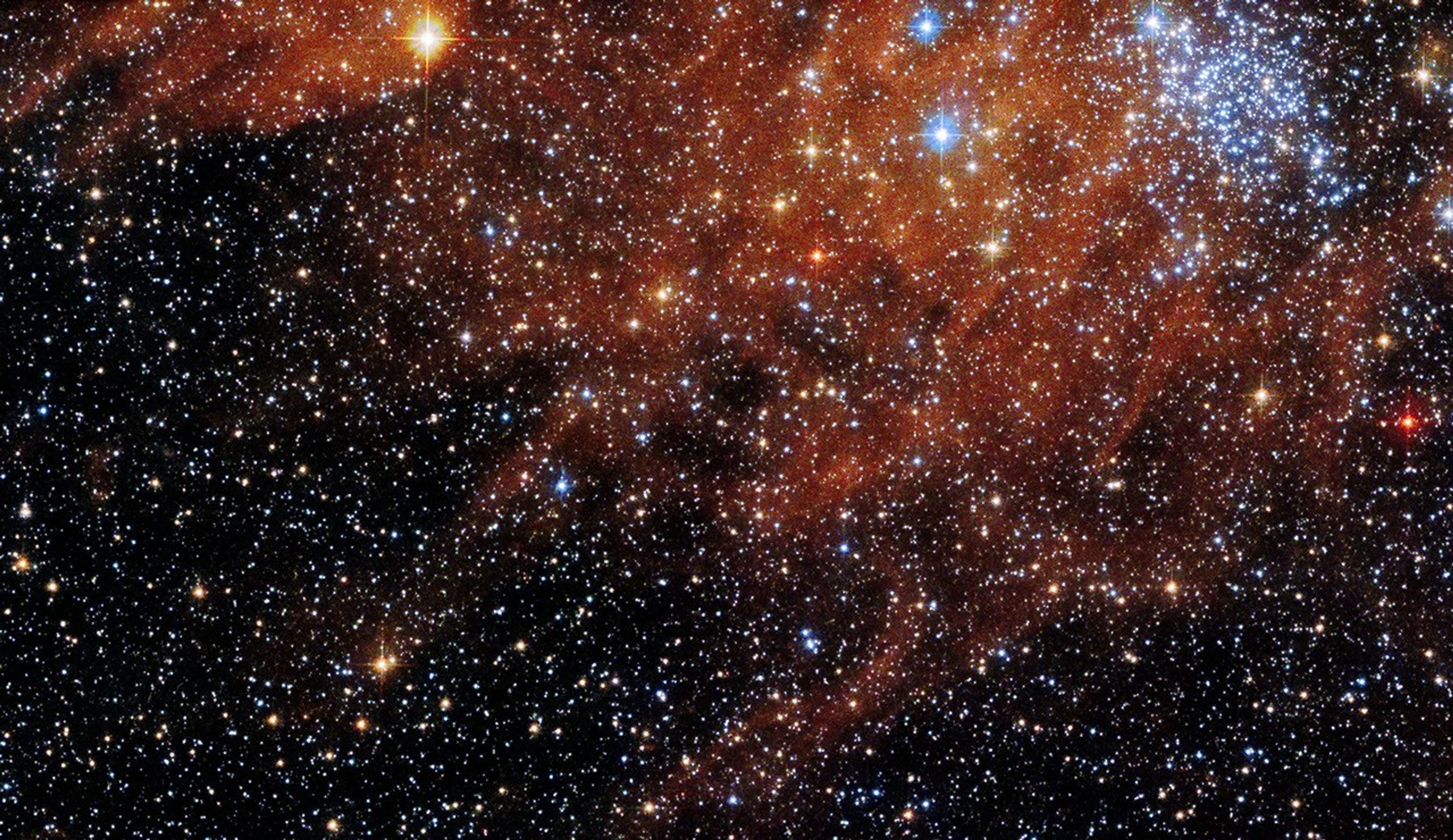 A thick grouping of blue-white stars in the upper right, with red-orange gas and dust in the upper half of the image. Thousands of stars dot the background throughout.
