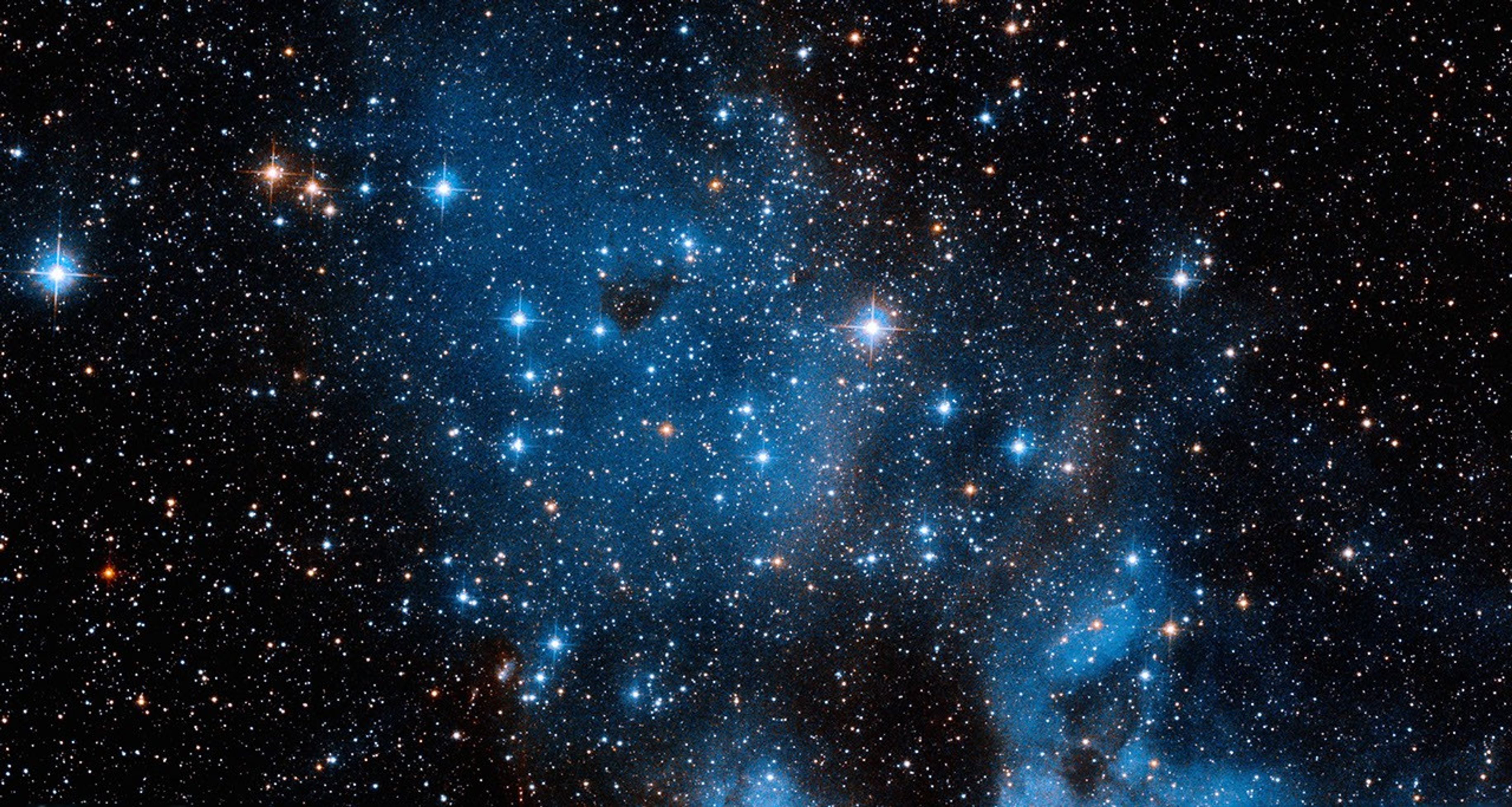 A couple dozen blue-white stars glow in the foreground, while a blue cloud of dust and thousands more distant stars shine in the background.