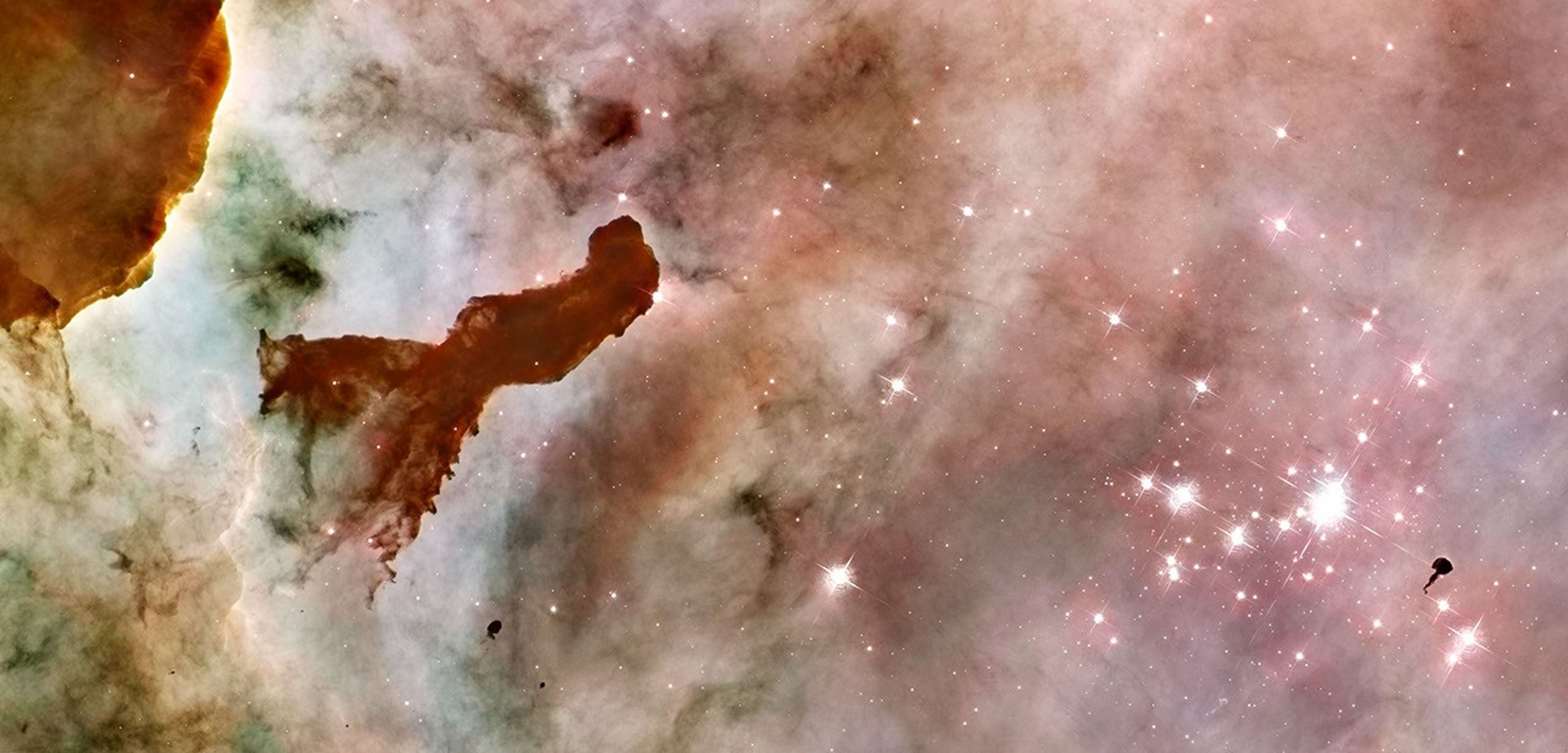On the left is a wall of yellow, orange, and green gas and dust, with a dark pink/red finger-like structure of gas. The background for the right half of the image is hazy pink dust, with a star cluster of a few dozen stars glowing on the right.