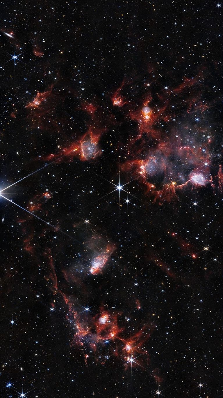 Tendrils and filaments of dust glow red in this infrared image. The region is bright with myriad stars, star clusters, and background galaxies. The dust is concentrated in the upper right and middle of the image, as well as the lower left.