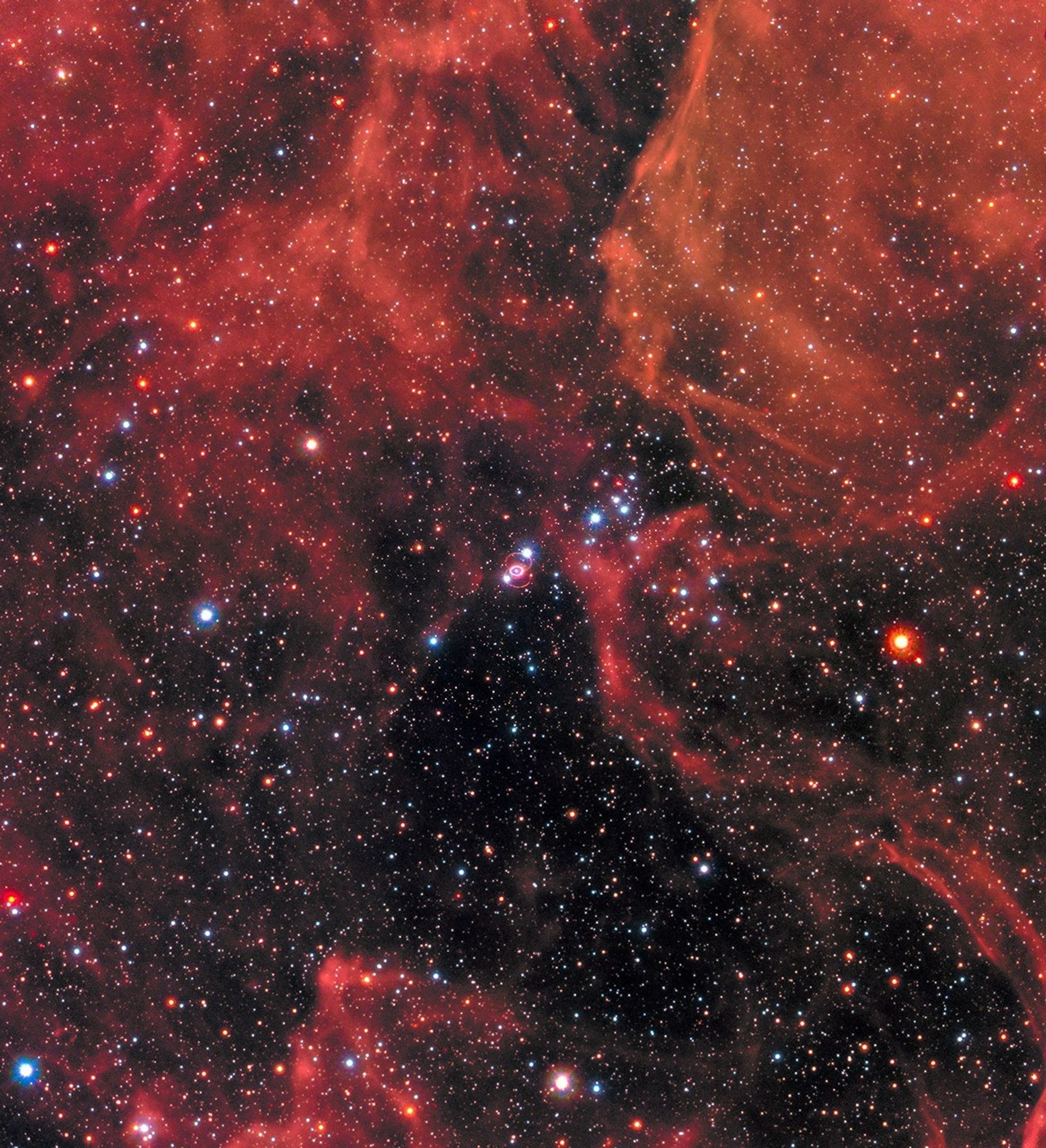 A black background is speckled with stars and red gas clouds. At image center is SN1987a with a series of bright rings circling it.