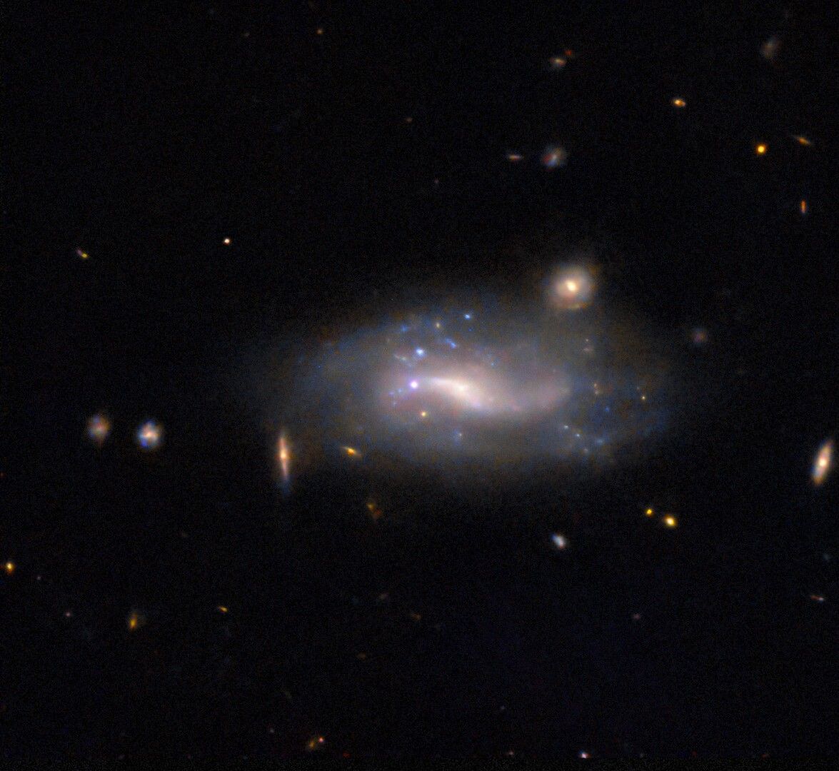A supernova appears as a small but bright pink-white dot at the exact center of the image. It lies in a spiral galaxy, close to the glowing center and next to bright patches of blue stars in the galaxy. A few minor galaxies are visible around the comparatively large spiral as small glowing disks, while distant galaxies appear as mere orangish spots and smudges, all are on a black background.