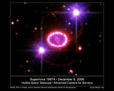 Una cadena de perlas rosáceas rodea una estrella explosionada. Fondo negro salpicado de estrellas. Dos estrellas brillantes, con rayos de difracción, en la parte inferior izquierda y superior derecha del anillo.