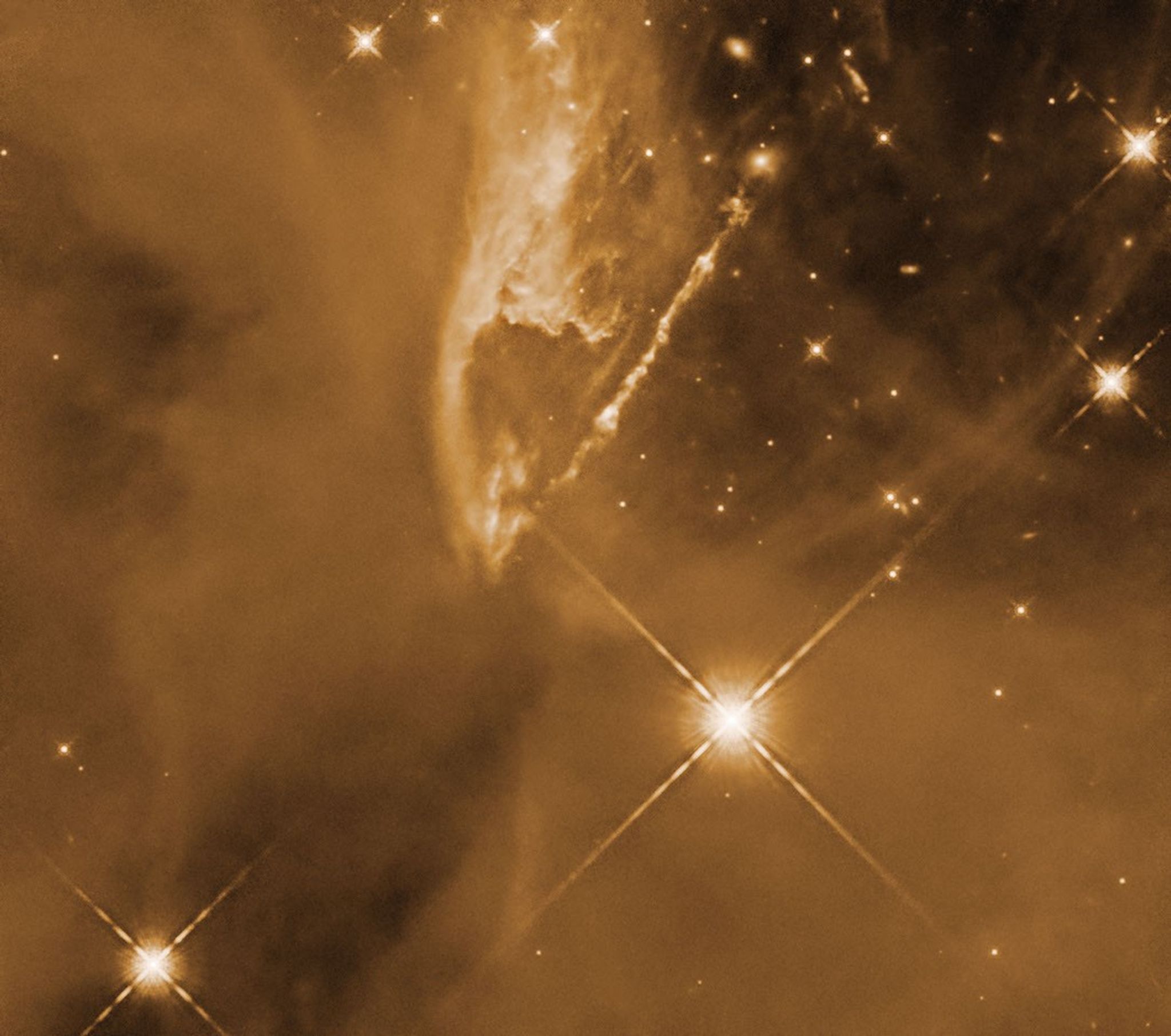A jet streams diagonally out from upper right corner of the orange-colored image to the center, across a field of gas clouds and stars. While most of the image is full of gas, the upper right corner is mostly clear of gas and dust, and more stars are visible in this darker space.