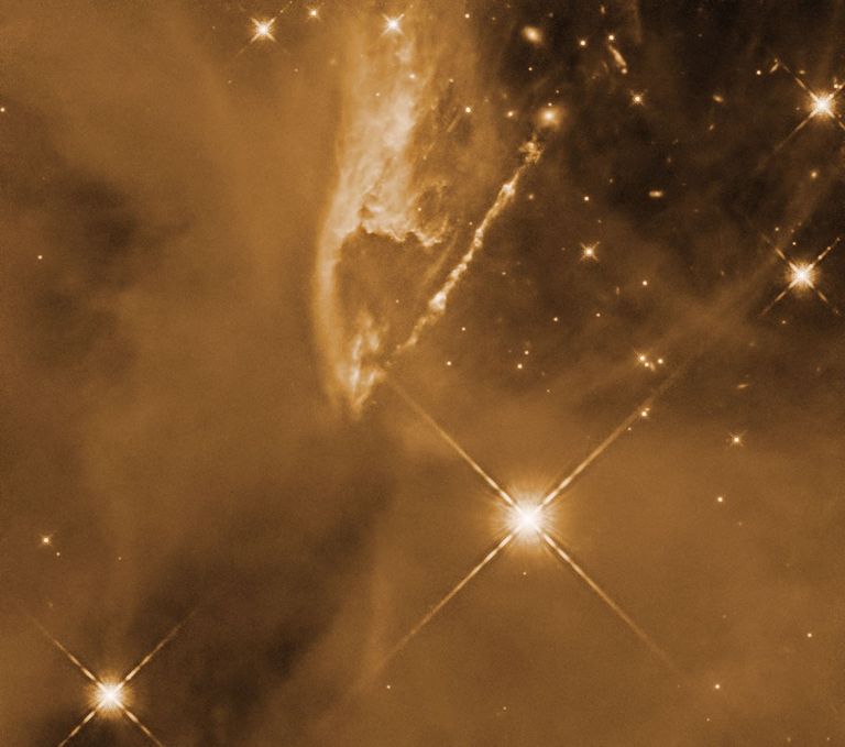 A jet streams diagonally out from upper right corner of the orange-colored image to the center, across a field of gas clouds and stars. While most of the image is full of gas, the upper right corner is mostly clear of gas and dust, and more stars are visible in this darker space.