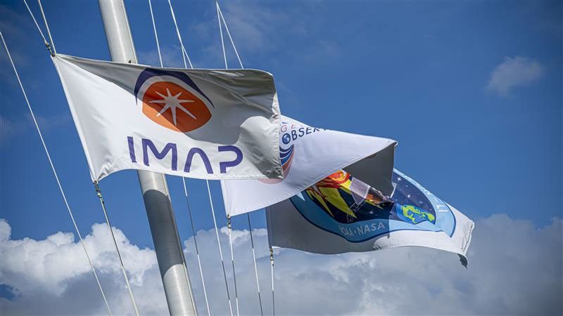 Flags for NASA’s IMAP (Interstellar Mapping and Acceleration Probe) mission and its two rideshares, NASA’s exosphere-studying Carruthers Geocorona Observatory and National Oceanic and Atmospheric Administration’s (NOAA) Space Weather Follow On–Lagrange 1 (SWFO-L1) spacecraft fly outside Astrotech Space Operations Facility near NASA’s Kennedy Space Center in Florida.