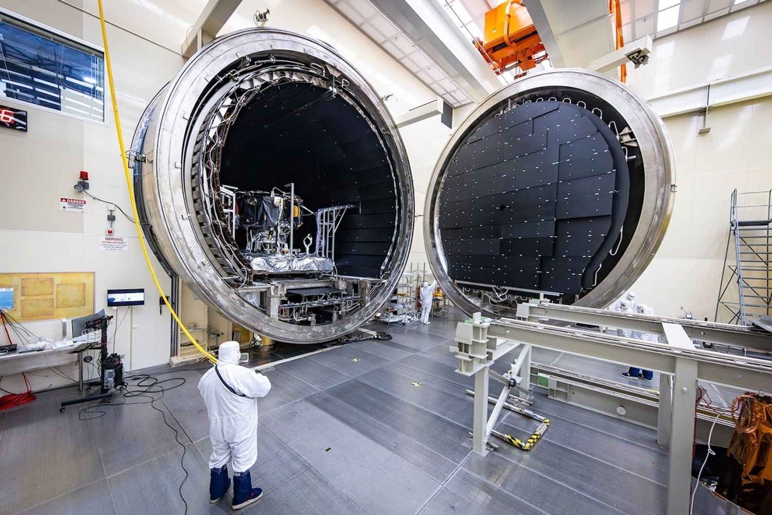 In a large laboratory, a circular door is open, revealing a large instrument inside a circular chamber. A person in a white full body suit stands outside.