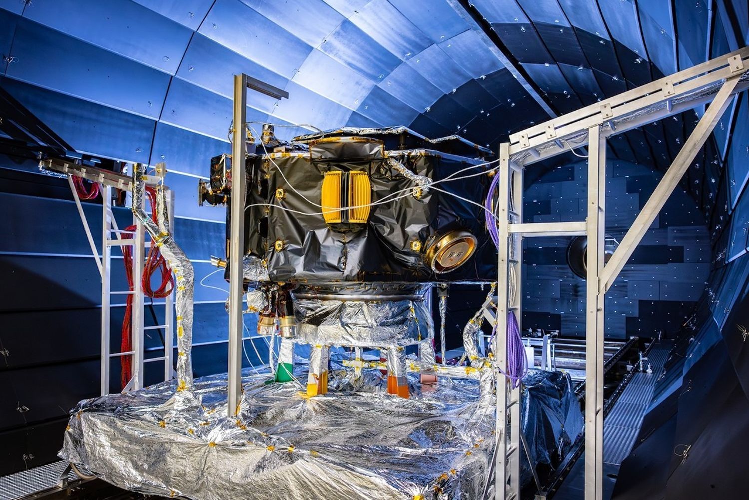 A large instrument with golden instruments wrapped in black material sits in a cylindrical paneled chamber atop a platform wrapped in silvery foil.