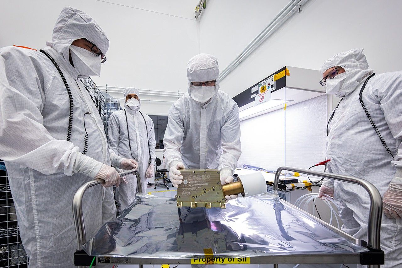 Four people dressed in white clean-room garmets stand around a gold-colored square instrument on a metal cart.