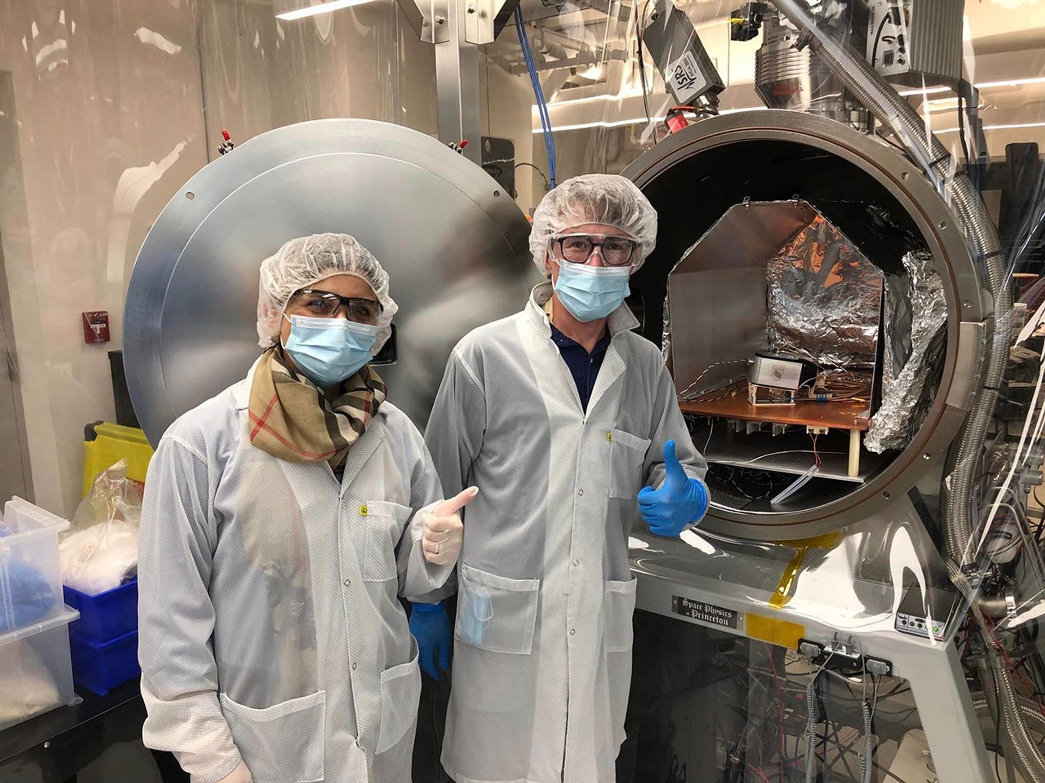 Two people wearing blue masks, white lab coats, and white hair nets have their thumbs up standing in front of a silver device used in calibration for the IMAP-Lo instrument.