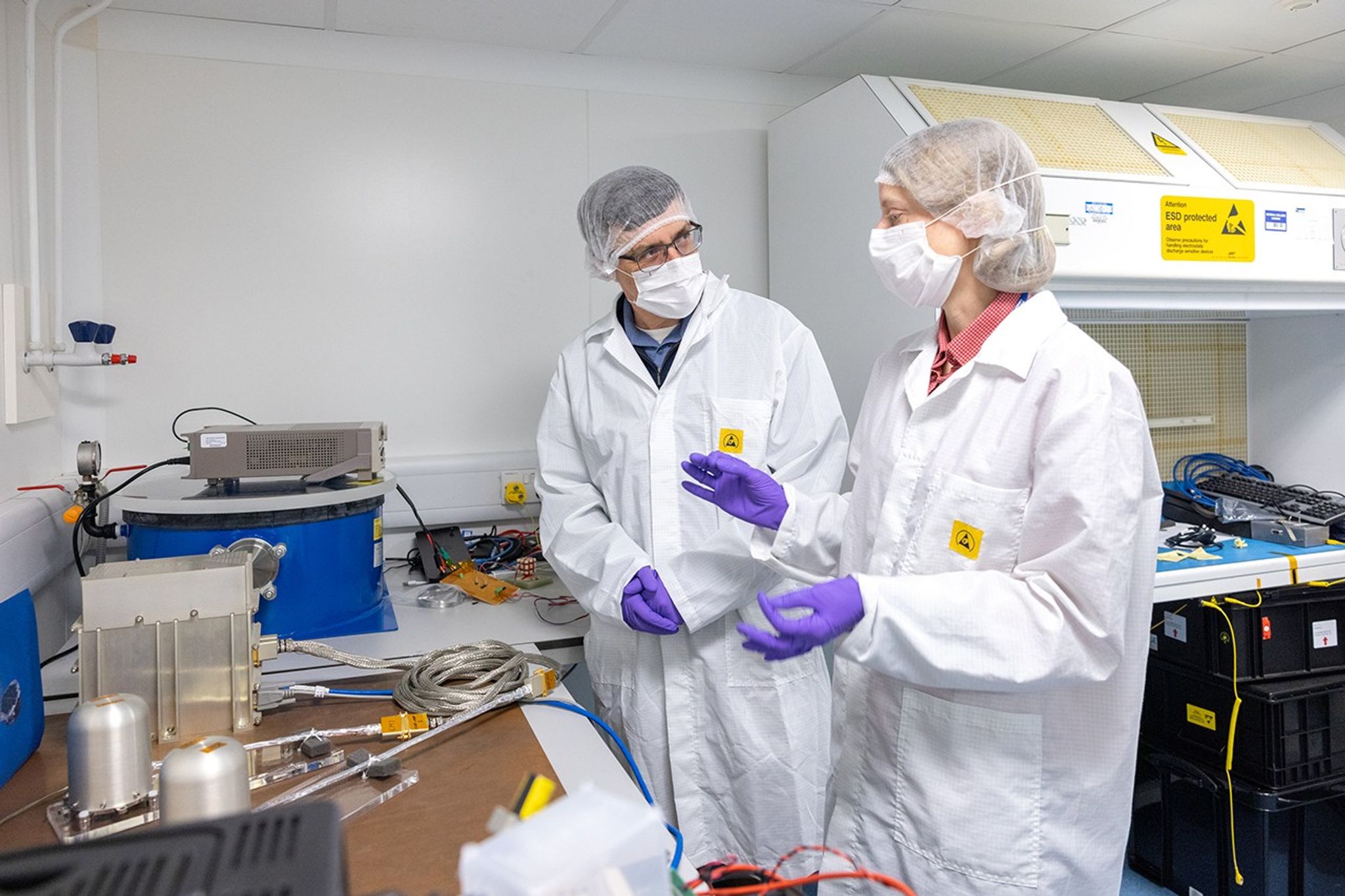 Two people wearing white lab coats, white hair nets, and purple gloves stand in front of the silver MAG instrument in a white clean room.
