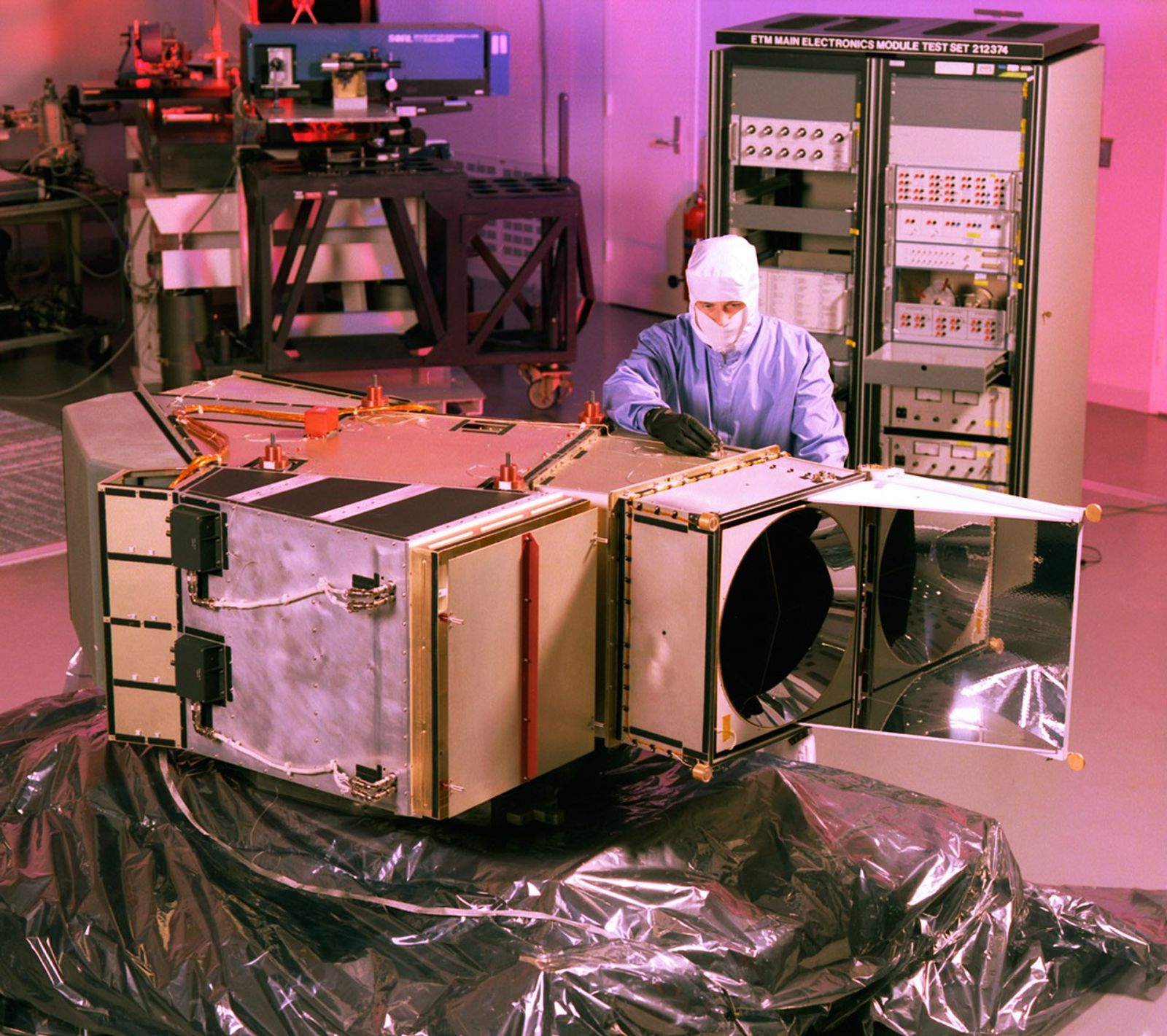 Landsat 7 ETM+ undergoing precision testing and calibration procedures in a controlled cleanroom environment, with technicians performing critical pre-flight verification of optical and mechanical systems.