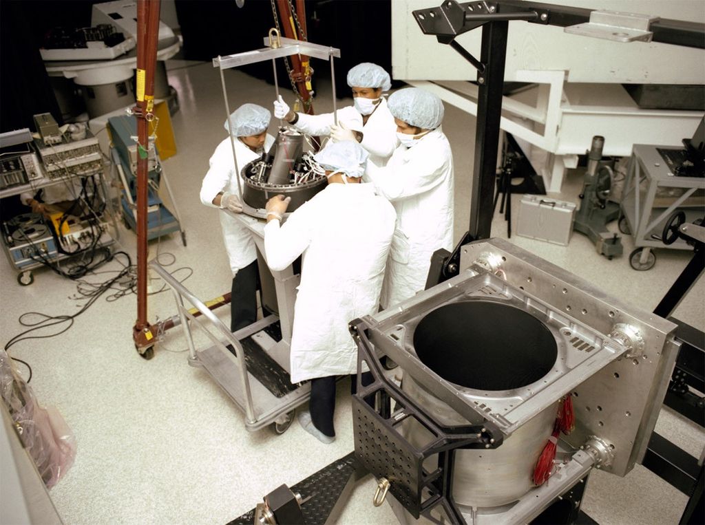 Four clean room technicians working together to install a Landsat 4 instrument component using lifting equipment.