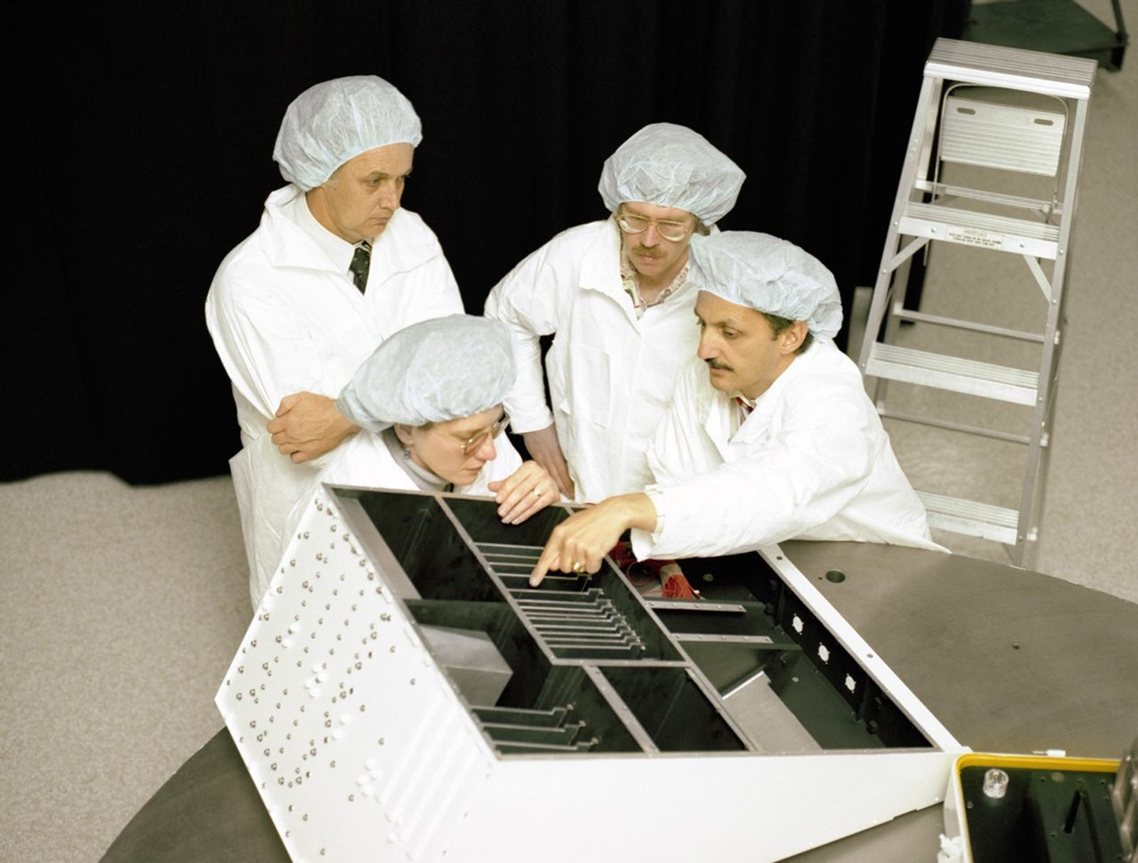 Engineers in clean room garments examining internal components of a Landsat 4 instrument during assembly and testing phase of the satellite development.