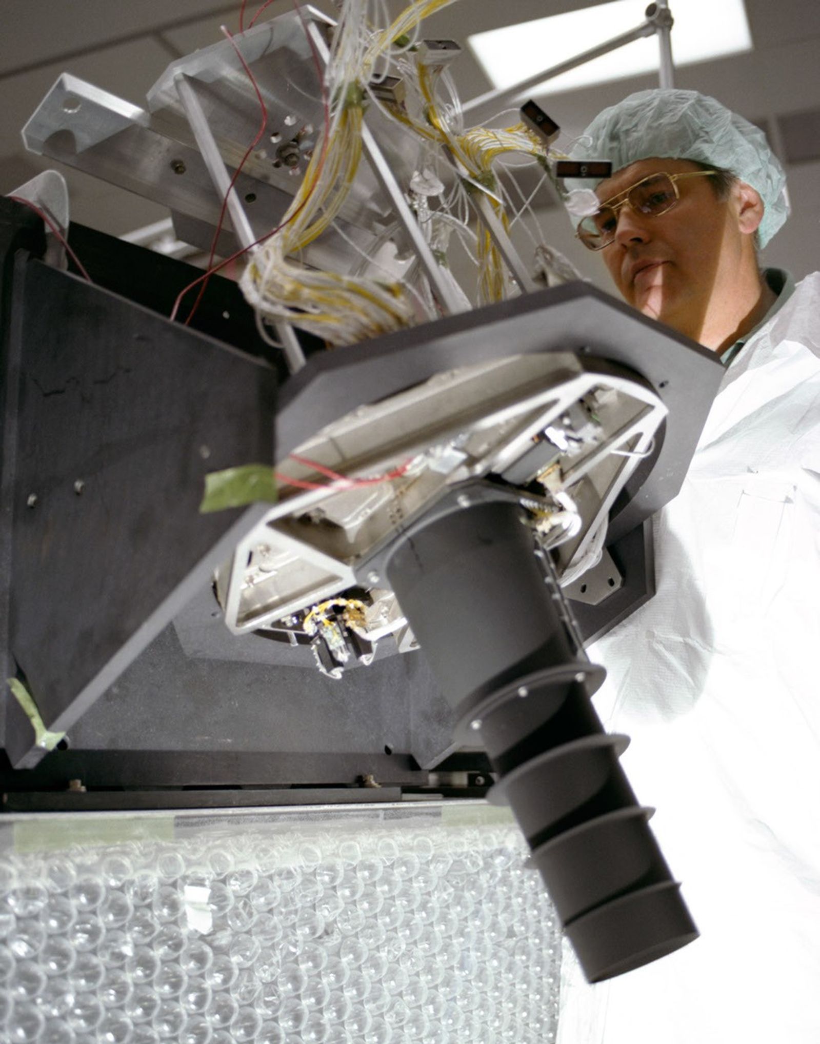Technician in clean room gear examining a Landsat 4 instrument assembly with exposed wiring and optical components.