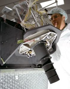 Technician in clean room gear examining a Landsat 4 instrument assembly with exposed wiring and optical components.
