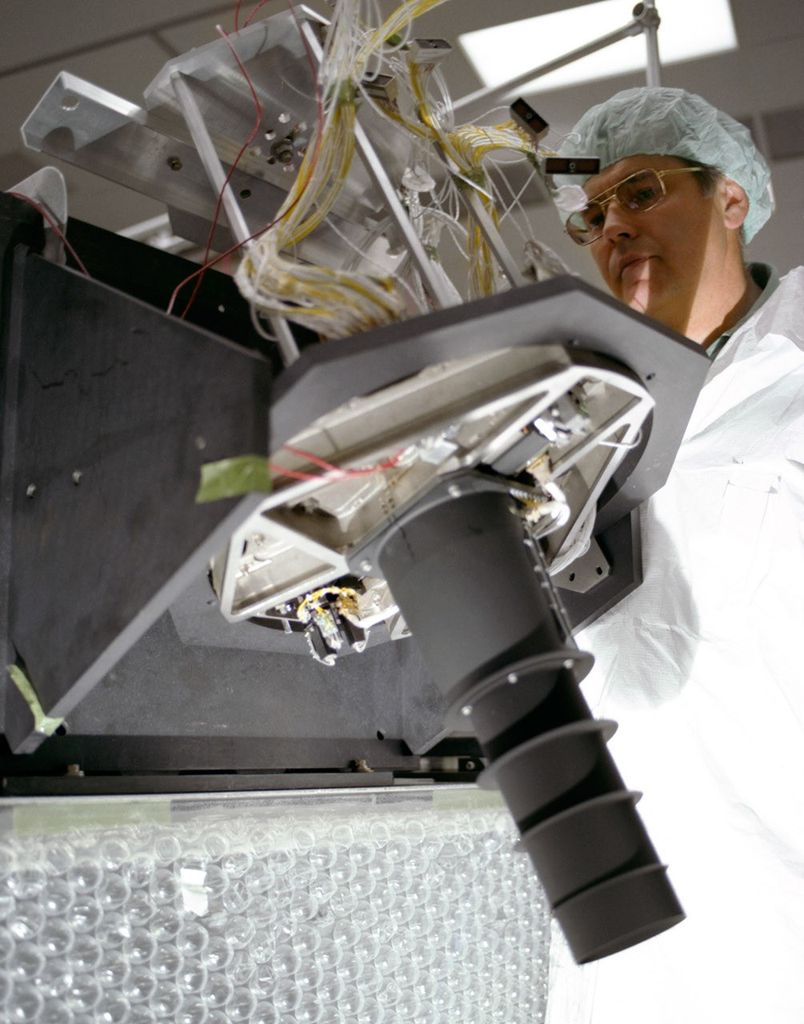 Technician in clean room gear examining a Landsat 4 instrument assembly with exposed wiring and optical components.
