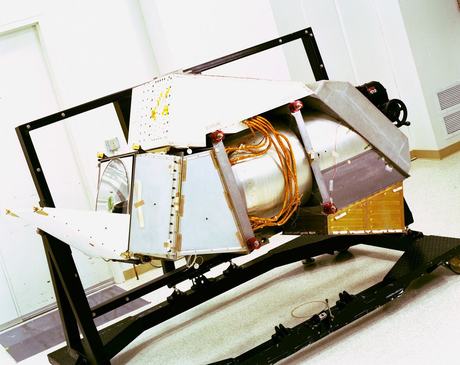 Landsat 4's Thematic Mapper instrument mounted on a test stand in a clean room, showing its metallic housing with exposed internal components and orange wiring harnesses.