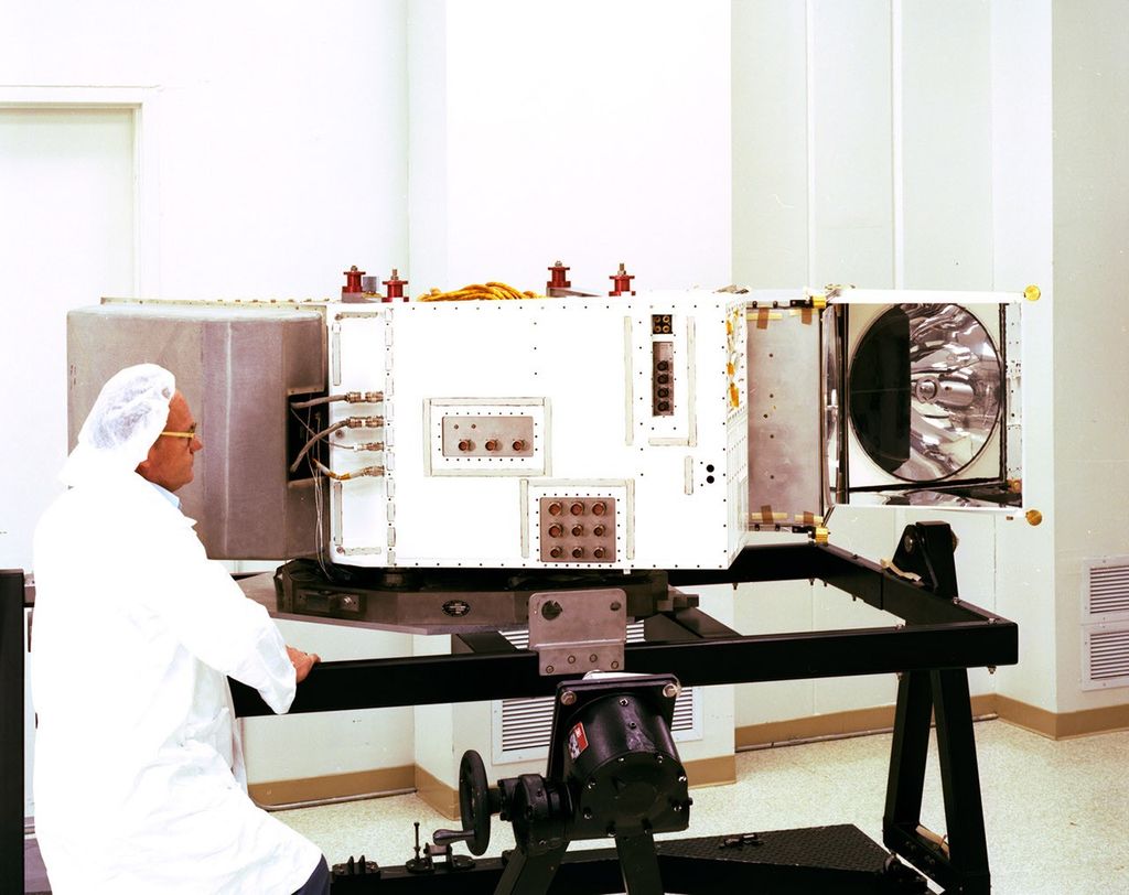 Clean room technician inspecting the Landsat 4 Thematic Mapper on a test stand, with the instrument housing open showing internal mirror assembly.