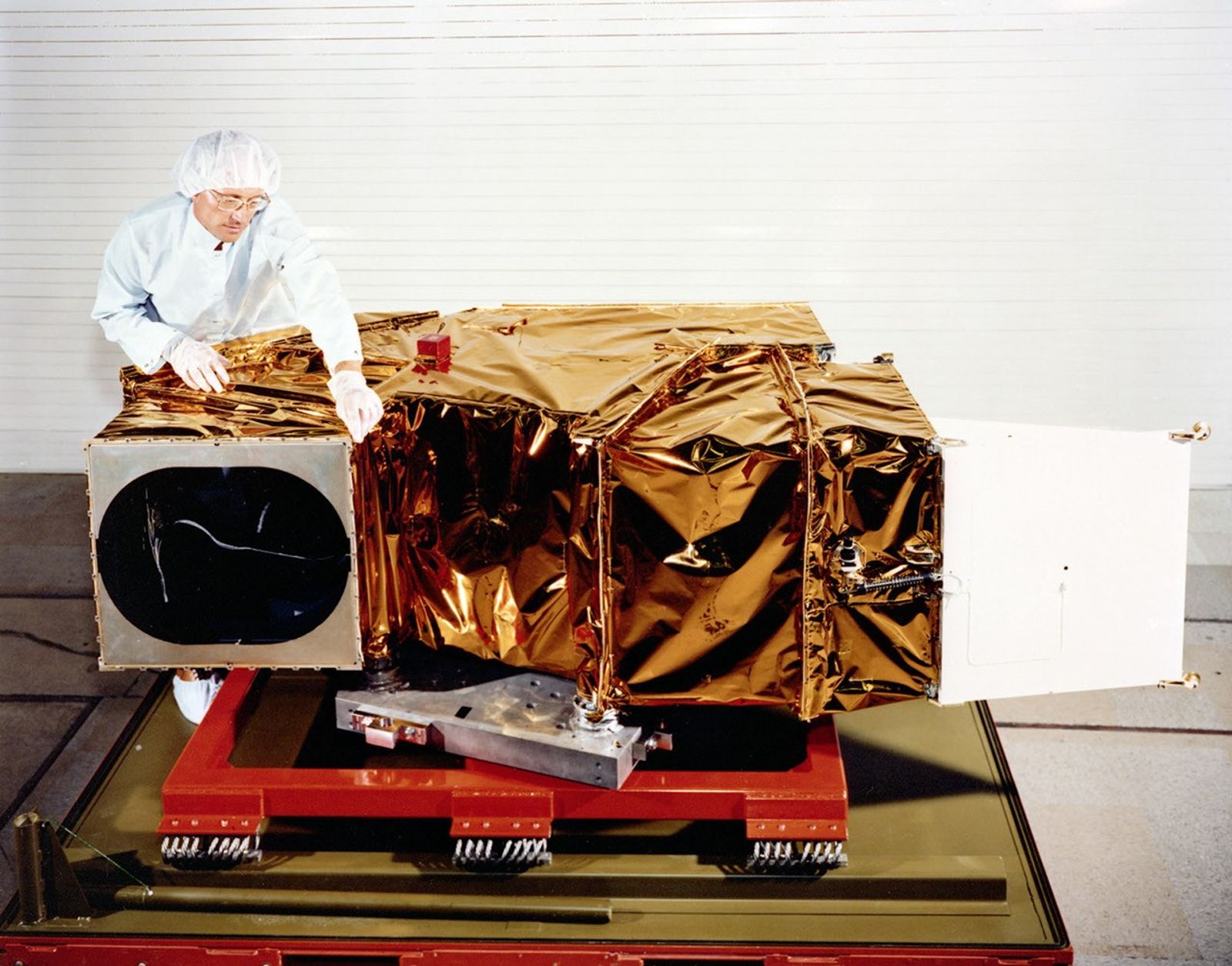 Clean room technician working on Landsat 4 Thematic Mapper instrument covered in gold thermal blankets with visible circular aperture and white panels on red platform.