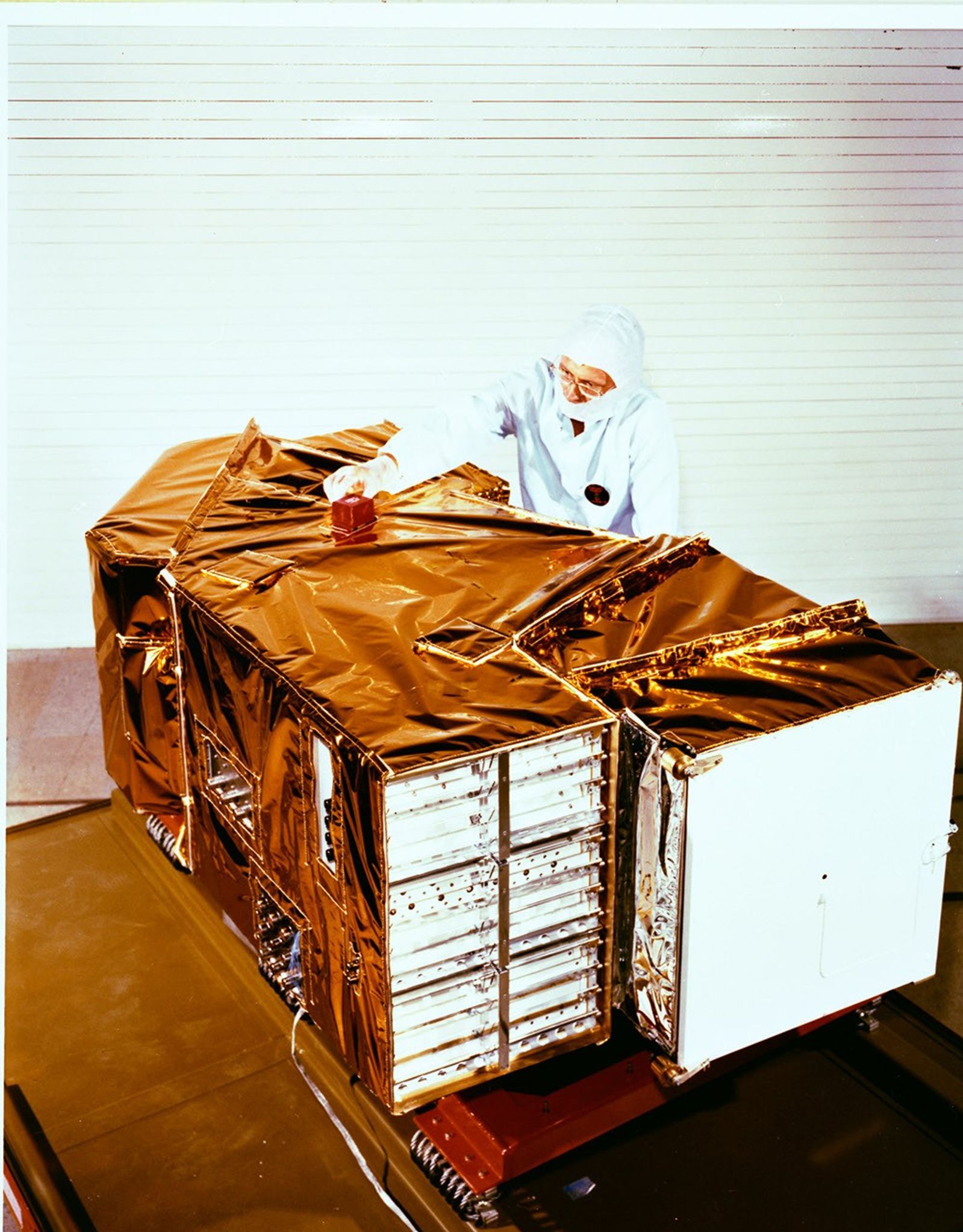 Landsat 4 Thematic Mapper instrument wrapped in gold thermal blankets with a technician in clean room gear working on top.