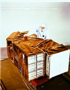 Landsat 4 Thematic Mapper instrument wrapped in gold thermal blankets with a technician in clean room gear working on top.