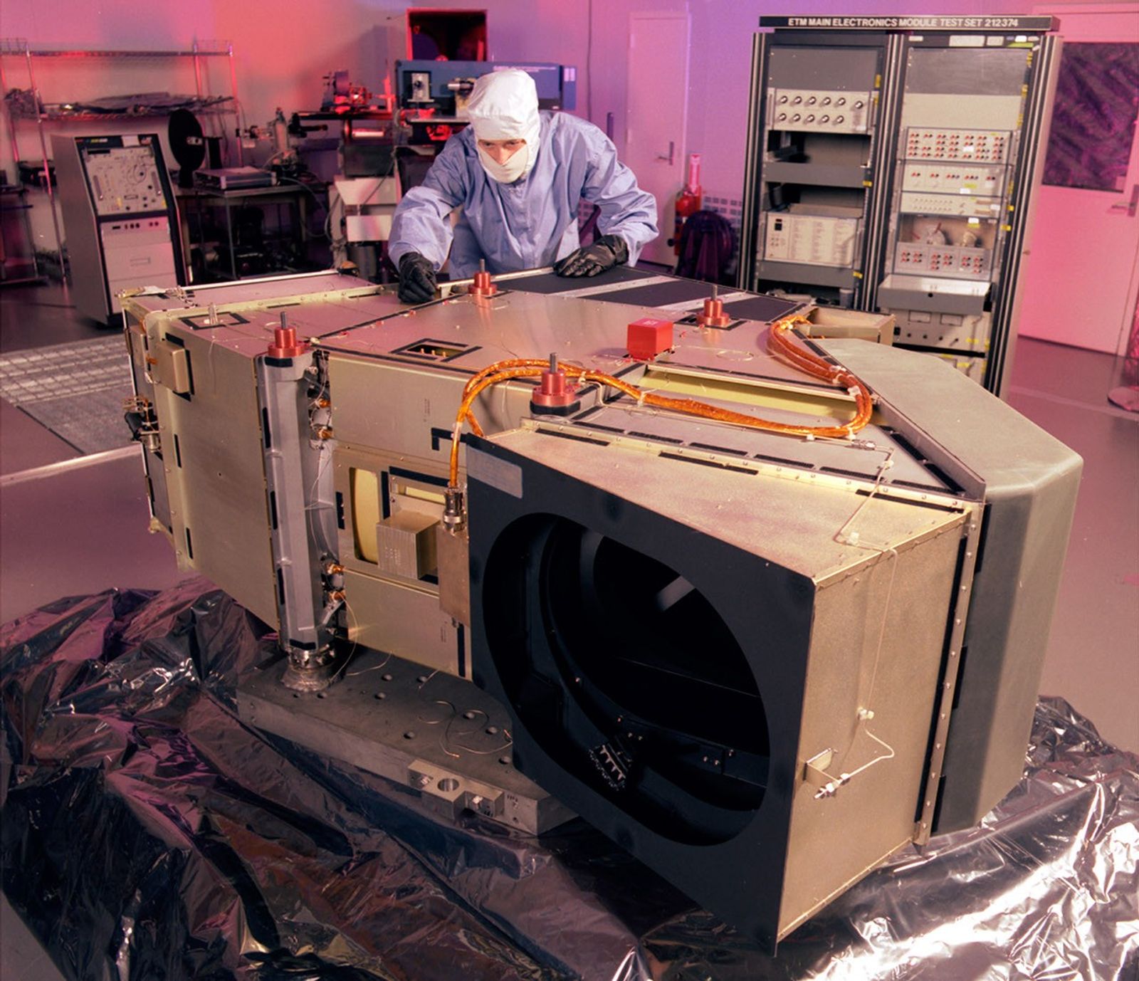 Landsat 7 ETM+ undergoing precision testing and calibration procedures in a controlled cleanroom environment, with technicians performing critical pre-flight verification of optical and mechanical systems.