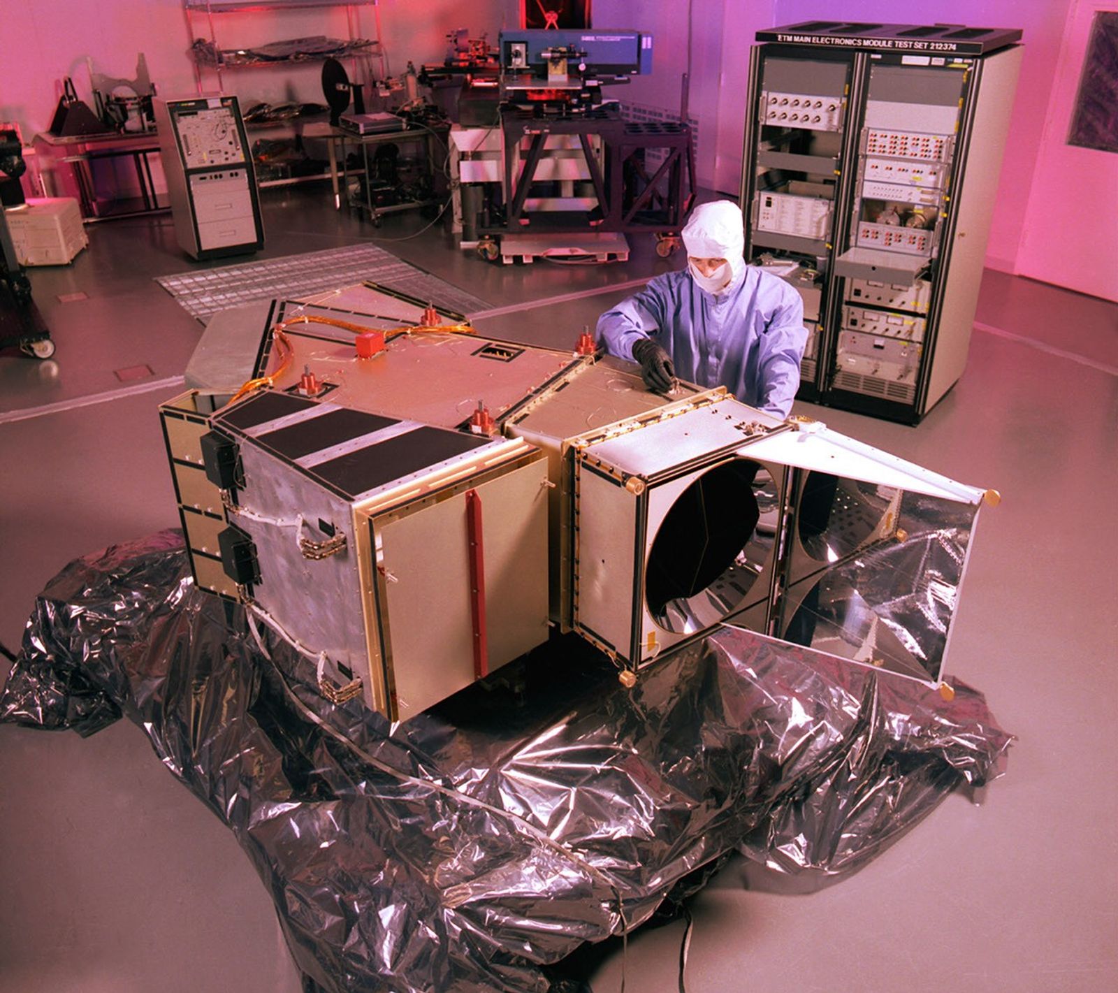 Landsat 7 ETM+ undergoing precision testing and calibration procedures in a controlled cleanroom environment, with technicians performing critical pre-flight verification of optical and mechanical systems.