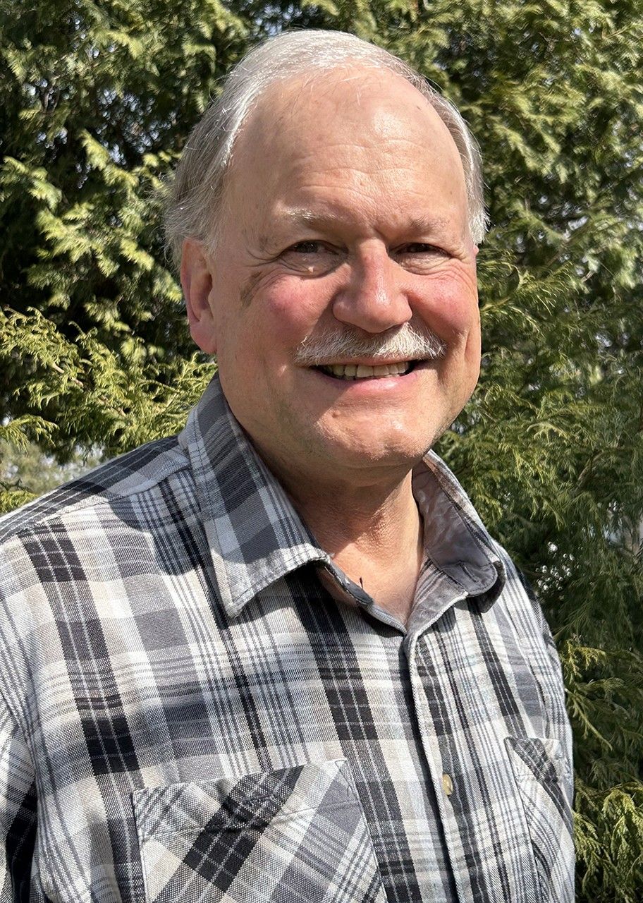 Brian Markham wears a black, white, and grey flannel shirt and stands in front of green vegetation.