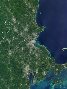 A natural-color satellite image of the Boston area. In the image, the majority of the left half is green with forests, urban areas like cities appear white, and the water of the Atlantic Ocean appears dark blue.