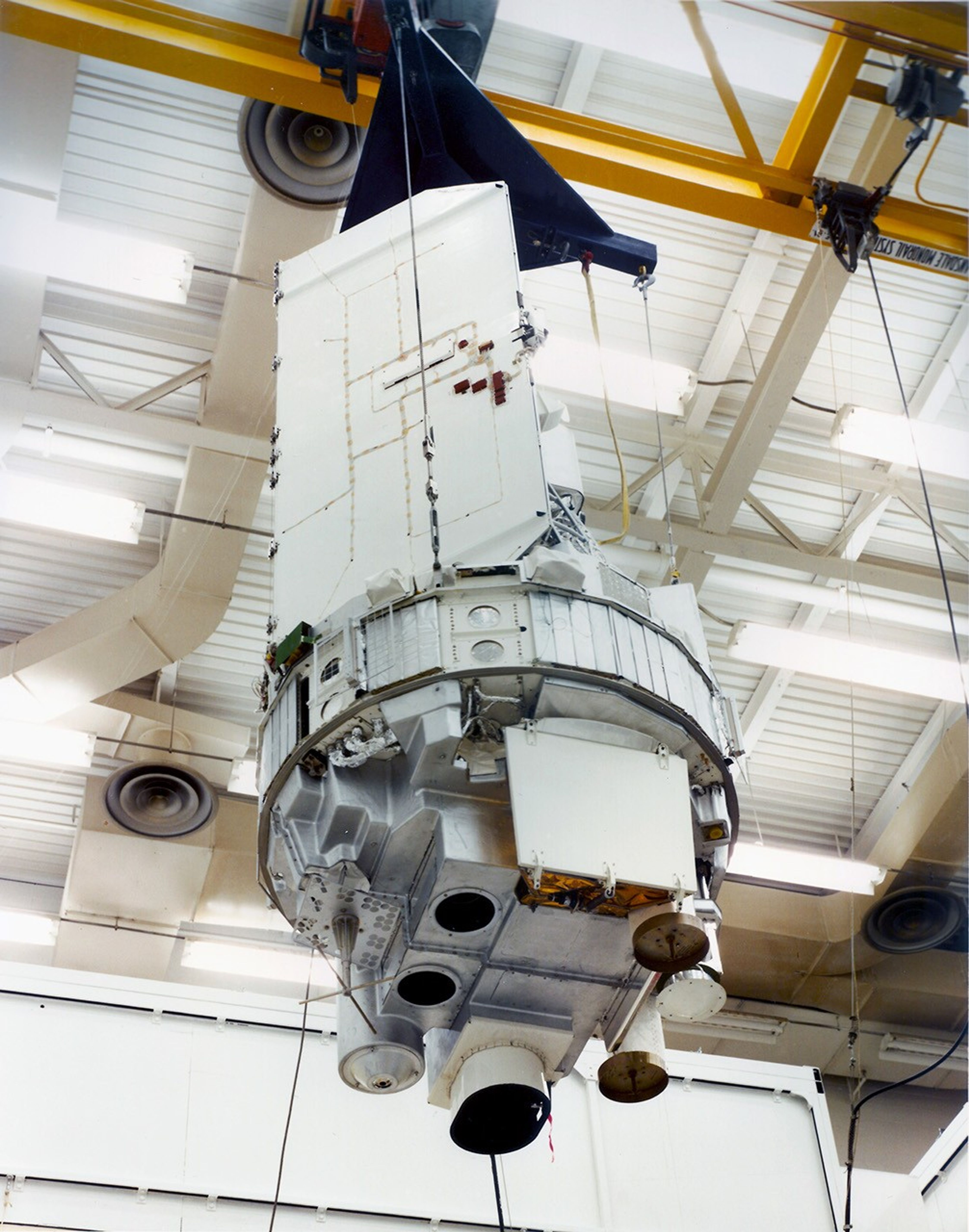 A photograph of the Landsat 3 satellite from below, hanging in a giant warehouse. The censors onboard Landsat 3 are visible in the undercarriage of this satellite.