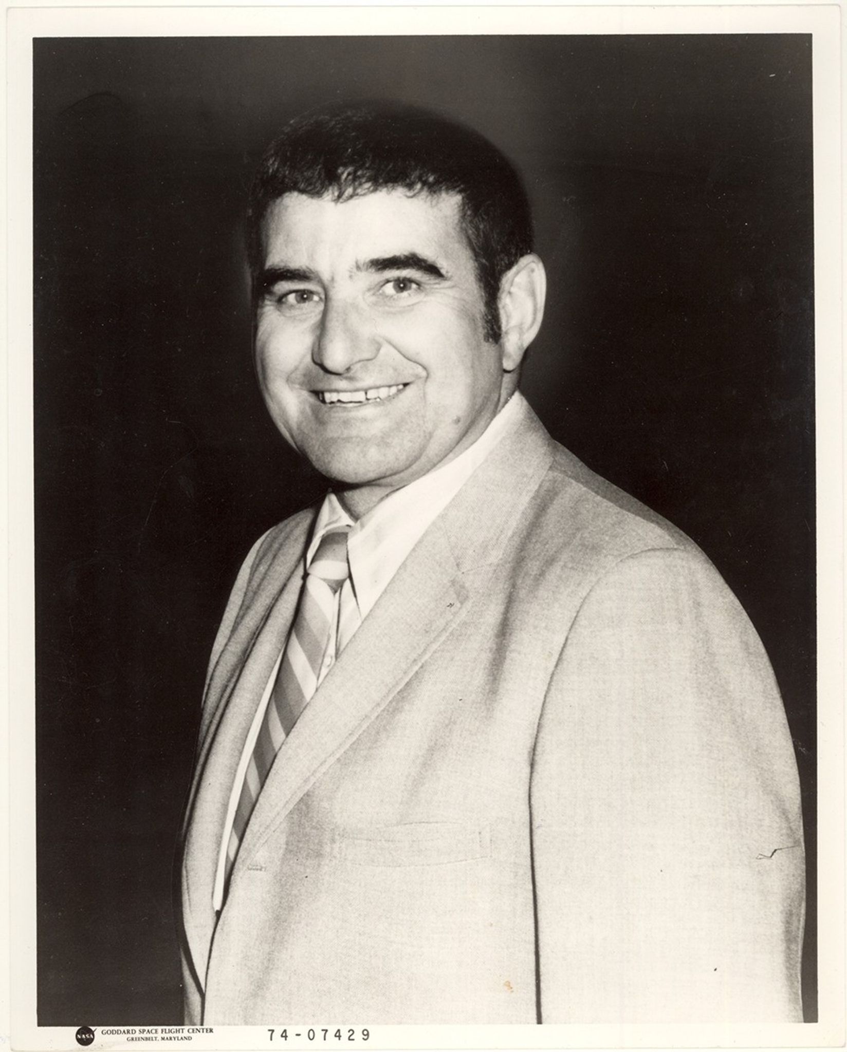 A black and white portrait photograph of a man in a light-colored suit and striped tie, smiling at the camera against a dark background. The image has a Goddard Space Flight Center photo identification number 74-07429 in the lower corner.