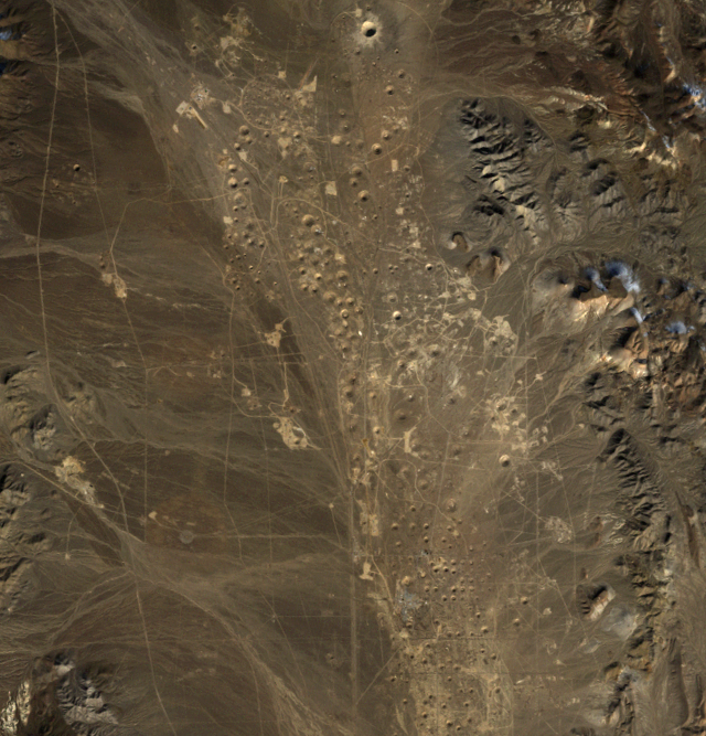 Aerial satellite image of Yucca Flat in Nevada showing numerous circular subsidence craters scattered across an arid desert landscape. The craters appear as distinct circular depressions against the brown desert terrain, with mountainous features visible on the right edge of the image.