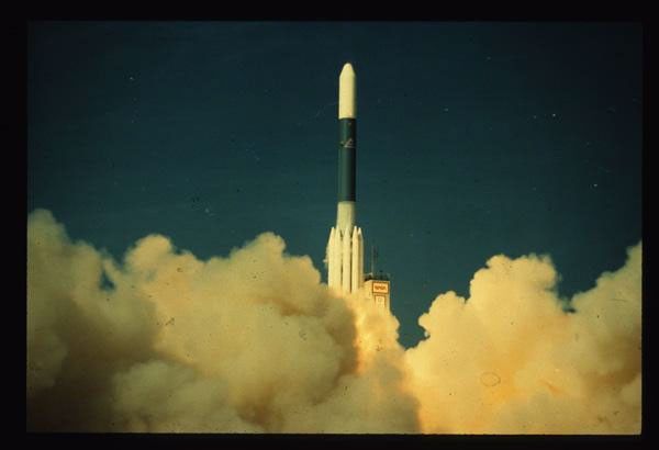 Landsat 4 satellite launching on Delta rocket with white and green stages, surrounded by launch exhaust clouds against blue sky.